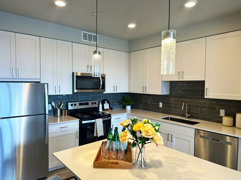 A modern kitchen here at Experience at Epicenter featuring white cabinetry, stainless steel appliances, a large island with quartz countertops, pendant lighting, and wood-style flooring.