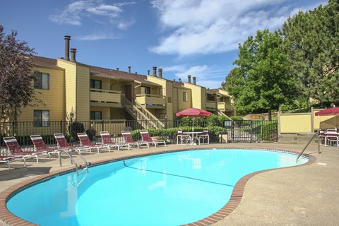 A sunny outdoor pool area here at Grand View featuring a curved swimming pool with clear blue water, brick-accented pool decking, red lounge chairs arranged along the fence, shaded patio tables with umbrellas, and landscaped greenery set in front of classic two-story apartment buildings under a bright blue sky.