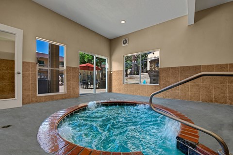 A relaxing indoor spa area here at Grand View featuring a circular bubbling hot tub with brick trim, clear blue water, and a metal handrail, surrounded by neutral tile flooring, large windows overlooking the outdoor patio, and warm beige walls that create a calm, comfortable retreat for residents year-round.