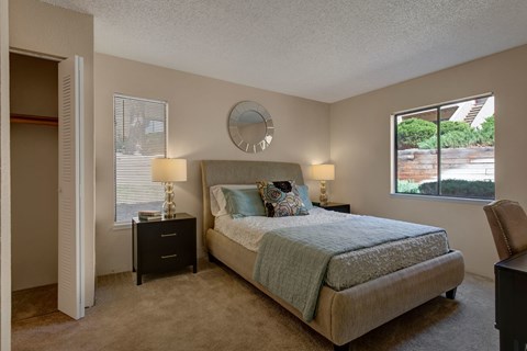 A bright and serene bedroom here at Grand View featuring a neatly made bed with soft blue and white bedding, a cushioned upholstered headboard, matching black nightstands with warm table lamps, plush carpeted flooring, a spacious open closet, and two windows that bring in natural light and views of greenery.
