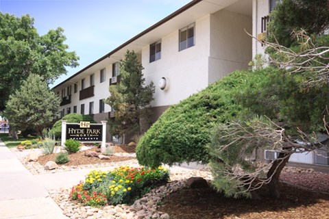 A landscaped community entrance here at Hyde Park North Apartments featuring a monument-style sign surrounded by colorful flowers, decorative rock beds, and mature trees. A multi-story apartment building with large windows sits behind the sign, creating a welcoming and well-maintained first impression of the community.