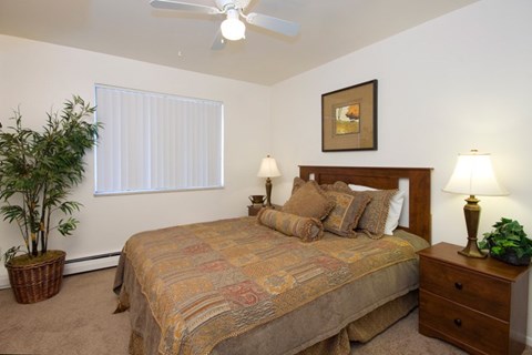 A comfortable bedroom here at Hyde Park North Apartments with beige carpet and light neutral walls, featuring a bed with patterned bedding, matching nightstands with lamps, and framed wall art above the headboard. A ceiling fan provides added comfort, while a window with vertical blinds and a potted plant create a calm, inviting atmosphere.