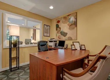 A welcoming leasing office here at Hyde Park Apartments with a wooden desk, guest chairs, and warm yellow walls. A large abstract art piece hangs above the desk, while a service window opens to an adjacent office area. Framed documents, soft lighting, and simple decor create a professional and inviting space for residents.