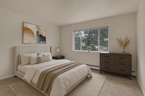 A bright, spacious bedroom here at Hyde Park Apartments with beige carpet, soft neutral walls, and a large window that brings in natural light and views of mature trees. The room features a comfortable bed with layered neutral bedding, a dresser, simple wall art, and a clean, calm layout that feels warm and inviting.
