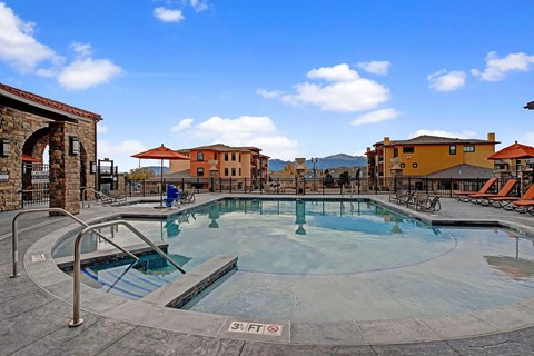 A scenic resort-style swimming pool here at La Bella Vita featuring clear water, built-in steps, and surrounding lounge chairs with umbrellas. A spacious stone pool deck overlooks Mediterranean-inspired buildings and distant mountain views, creating a relaxing outdoor amenity space for residents to enjoy year-round.