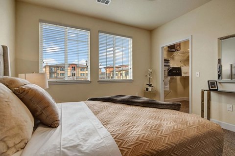 A serene primary bedroom here at La Bella Vita featuring a comfortable bed dressed in soft neutral bedding with layered pillows and a folded throw, positioned beside large windows with blinds that fill the room with natural light and views of the surrounding community. An open walk-in closet and simple accent furnishings complete the airy, inviting space.