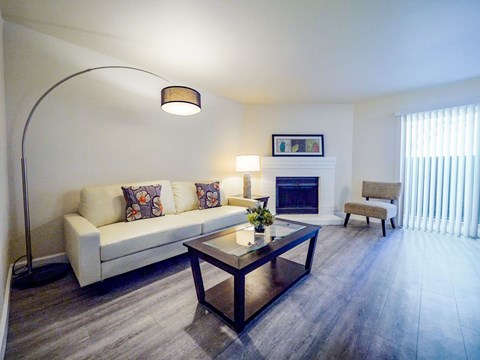 A bright living room here at Retreat at Austin Bluffs featuring a light-colored sofa with patterned accent pillows, a glass-top coffee table, and wide plank flooring. A curved floor lamp and table lamp add warm lighting, while a corner fireplace, sliding glass door with vertical blinds, and simple accent chair create a comfortable, open layout.