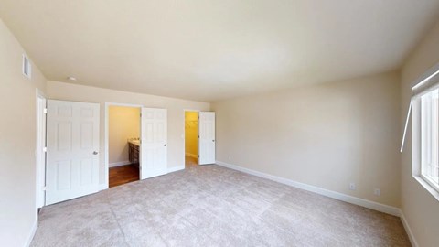 A spacious neutral bedroom here at Retreat at Austin Bluffs featuring soft carpet flooring, light-toned walls, and a wide layout with ample room for furnishings. Double doors open to an adjacent bathroom and walk-in closet, while a large window brings in natural light and creates a bright, comfortable atmosphere.