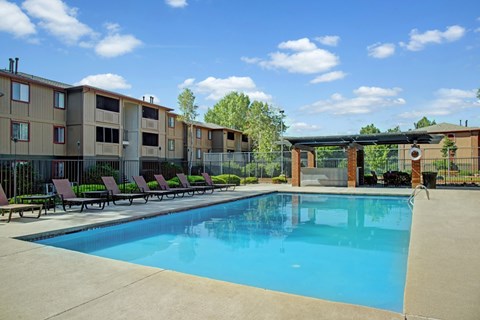 A refreshing outdoor swimming pool here at Retreat at Austin Bluffs featuring clear blue water surrounded by a spacious concrete deck with multiple lounge chairs for relaxing. The fenced pool area includes shaded seating under a pergola, with well-maintained apartment buildings and mature trees in the background beneath a bright blue sky.
