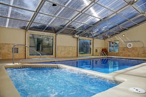A bright indoor swimming pool here at Retreat at Austin Bluffs featuring clear blue water beneath a translucent enclosed roof that allows natural light to fill the space. The pool area includes built-in steps, metal handrails, and a smooth surrounding deck, with large windows, warm-toned wall finishes, and a clean, inviting atmosphere.