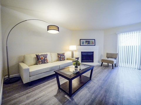 A bright living room here at Retreat at Austin Bluffs featuring a light-colored sofa with patterned accent pillows, a glass-top coffee table, and wide plank flooring. A curved floor lamp and table lamp add warm lighting, while a corner fireplace, sliding glass door with vertical blinds, and simple accent chair create a comfortable, open layout.