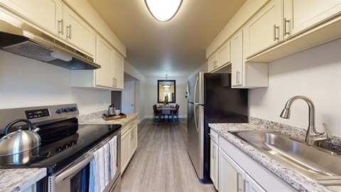 A bright galley-style kitchen here at Retreat at Austin Bluffs featuring white shaker-style cabinetry, speckled countertops, and wood-look flooring that extends toward a dining area. The space includes a refrigerator, range, sink, and modern lighting, creating a clean, functional layout with a warm, inviting feel.