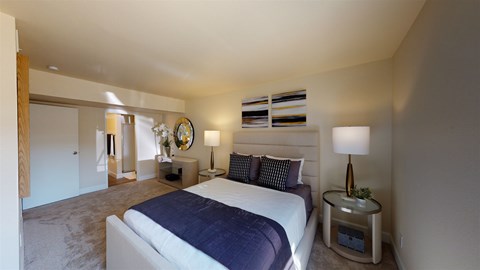 A bright primary bedroom here at Retreat at Austin Bluffs featuring a plush upholstered bed with layered neutral bedding, matching nightstands with modern lamps, and soft carpet flooring. The room includes abstract wall art, a dresser with mirror, and an open layout that connects toward an adjacent bathroom area, creating a calm and comfortable retreat.
