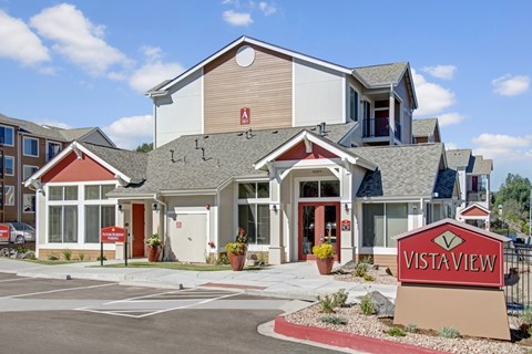 A welcoming exterior clubhouse building here at Vista View featuring peaked rooflines, neutral siding with red accent panels, large windows, and a clearly marked entrance, with landscaped planters, paved walkways, and a prominent Vista View monument sign set against a bright blue sky with scattered clouds.