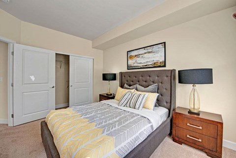 A cozy staged bedroom here at Vista View featuring a tufted upholstered bed with patterned neutral bedding, matching wood nightstands with modern lamps, soft carpet flooring, and sliding closet doors, complemented by warm wall tones and simple artwork that create a comfortable and inviting sleeping space.