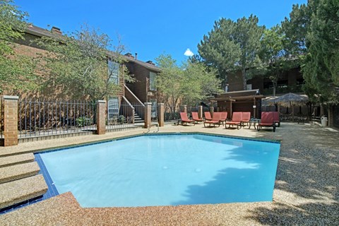 A sunny outdoor swimming pool here at Windtree featuring clear blue water, a textured pool deck, comfortable lounge chairs, shaded seating areas, mature trees, and surrounding brick apartment buildings that create a relaxed and inviting community setting.
