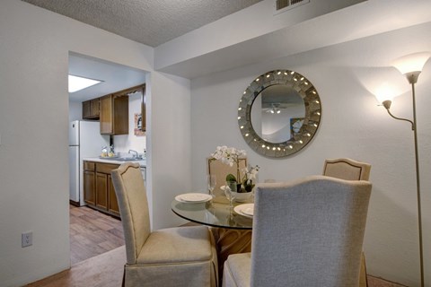 A warm and inviting dining area here at Windtree featuring a round glass-top dining table with upholstered chairs, soft carpet flooring, and a decorative wall mirror accented with subtle lighting, positioned just off the kitchen to create an open, comfortable space ideal for everyday meals and casual entertaining.