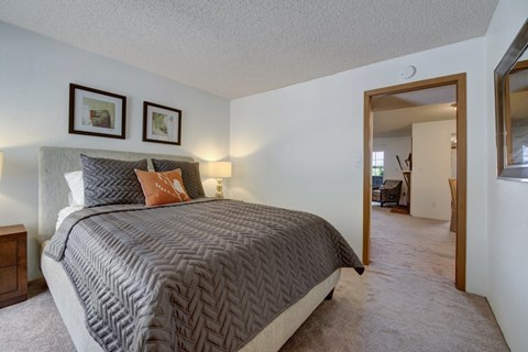 A cozy bedroom here at Windtree featuring plush carpeting, a neutral upholstered bed with textured gray bedding and accent pillows, warm bedside lighting, framed wall art above the headboard, and an open doorway leading into the adjoining living area, creating a comfortable and inviting private retreat.