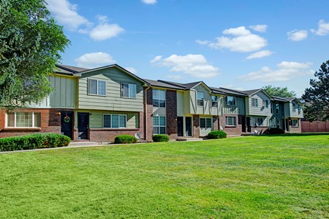 A bright exterior view here at Yorkshire Square featuring two-story townhome-style apartment buildings with brick and green siding accents, private front entry doors, large windows, neatly trimmed shrubs, and an expansive grassy courtyard under a blue sky that creates an open and welcoming residential setting.