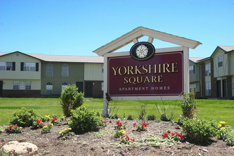 A welcoming community sign here at Yorkshire Square featuring a red monument sign with the property name set in landscaped greenery, colorful flowers, and trimmed shrubs, with two-story apartment buildings and a wide grassy courtyard visible in the background under a clear blue sky.