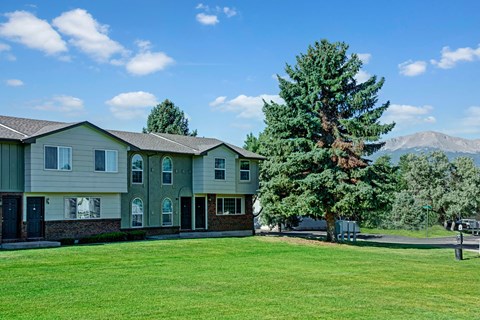 A spacious green courtyard here at Yorkshire Square featuring well-kept grass, mature evergreen trees, and a row of two-story apartment homes with neutral siding and brick accents, set beneath a bright blue sky with soft clouds and distant mountain views in the background.