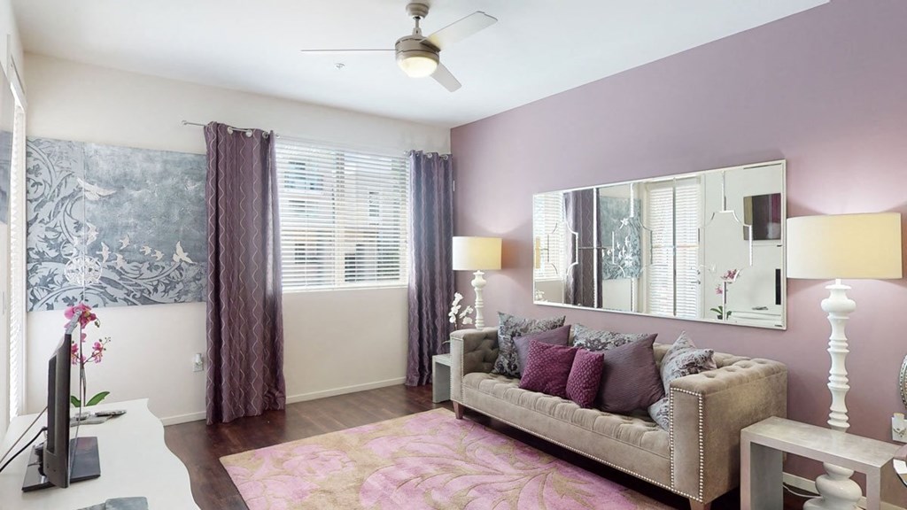 Modern living room here at Artisan at Main Street Metro Apartments with lavender walls, grey tufted sofa, and pink cushions. Large mirror, abstract art, and floral rug complement the chic decor.