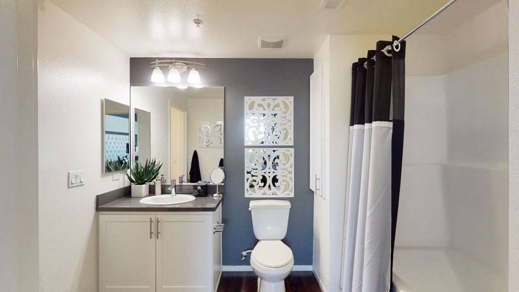 A modern bathroom here at Artisan at Main Street Metro Apartments with a sleek design features a white vanity, round basin, and rectangular mirror. There's a toilet, decorative wall art, and a shower with a black and white curtain. The space is bright and organized.
