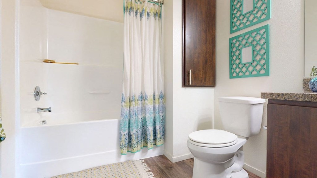 Bright bathroom here at Artisan at Main Street Metro Apartments with a white tub and shower, blue-patterned curtain, wood cabinet, white toilet, and turquoise wall decor. Cozy and clean atmosphere.