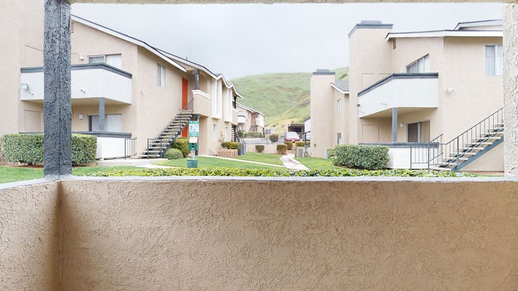 A view from a balcony overlooks a courtyard with beige Hillside Village Apartment Homes buildings. Green lawns and hedges line the pathways, leading to distant green hills.