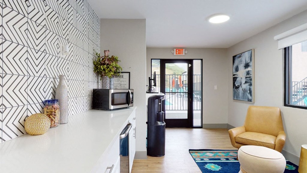 Modern room here at Hillside Village Apartment Homes with geometric patterned wall, countertop with a microwave and decor, sunny exit to pool area. Cozy seating with colorful rug. Calm ambiance.