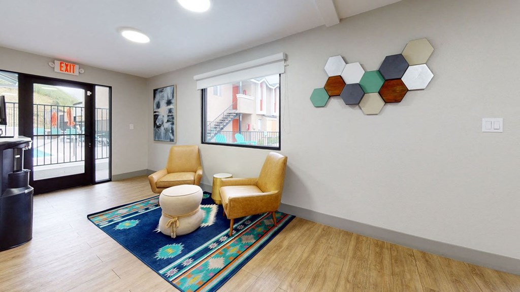 Cozy room here at Hillside Village Apartment Homes with two tan chairs, a patterned blue rug, and a round ottoman. Abstract wall art and hexagonal decor add color. Bright and inviting.