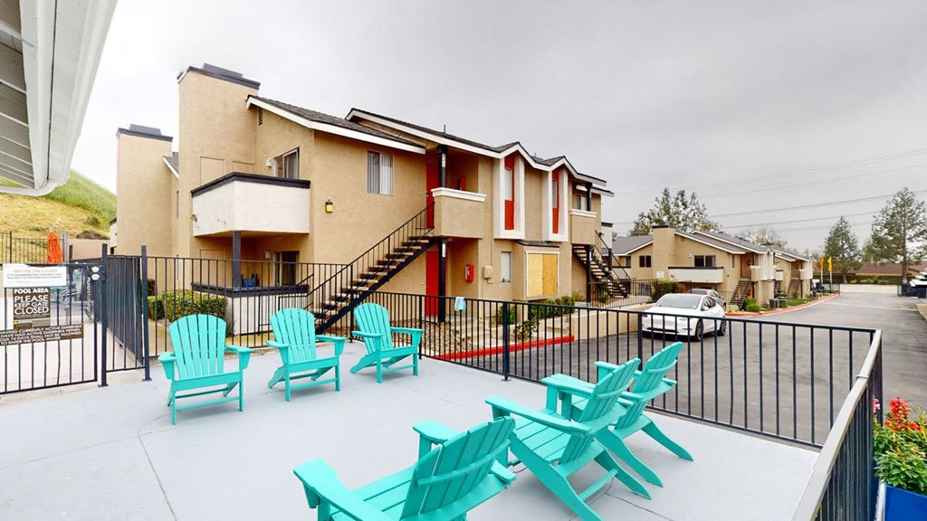 Two-story beige Hillside Village Apartment Homes buildings under a cloudy sky, with turquoise chairs on a patio in the foreground. A white car is parked on a paved lot.