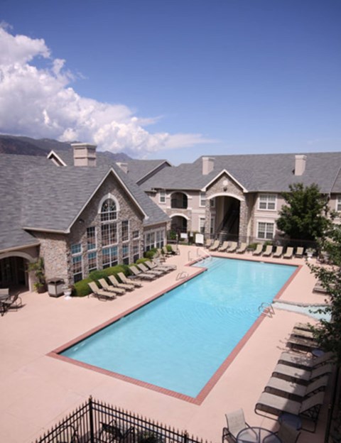 A resort-style outdoor pool here at Camelback Pointe featuring a long rectangular swimming pool surrounded by lounge chairs, light-colored pool decking, and elegant apartment buildings with stone accents, creating a relaxing community space under a bright sky with scenic mountain views in the background.
