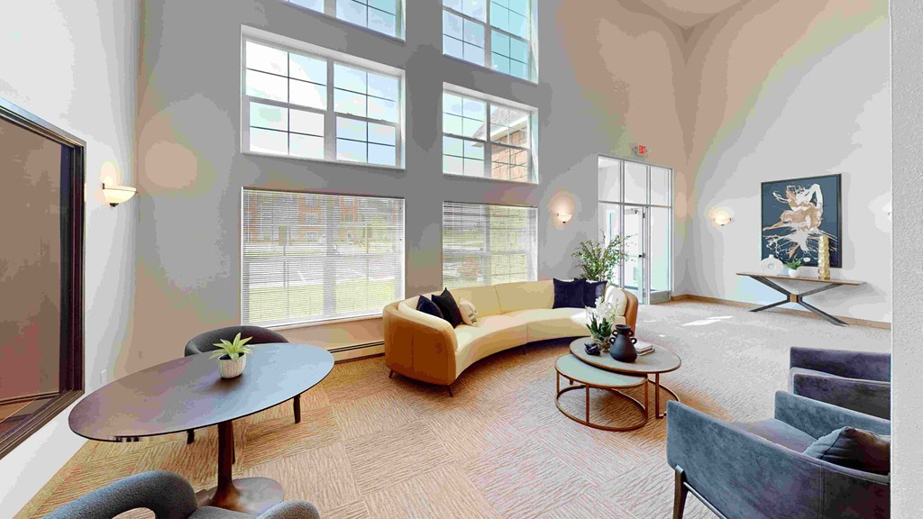 Spacious modern seating area here at The Club at Heritage Apartment Homes with large windows, curved beige sofa, dark round tables with plants, and artwork on walls. Bright and inviting atmosphere.
