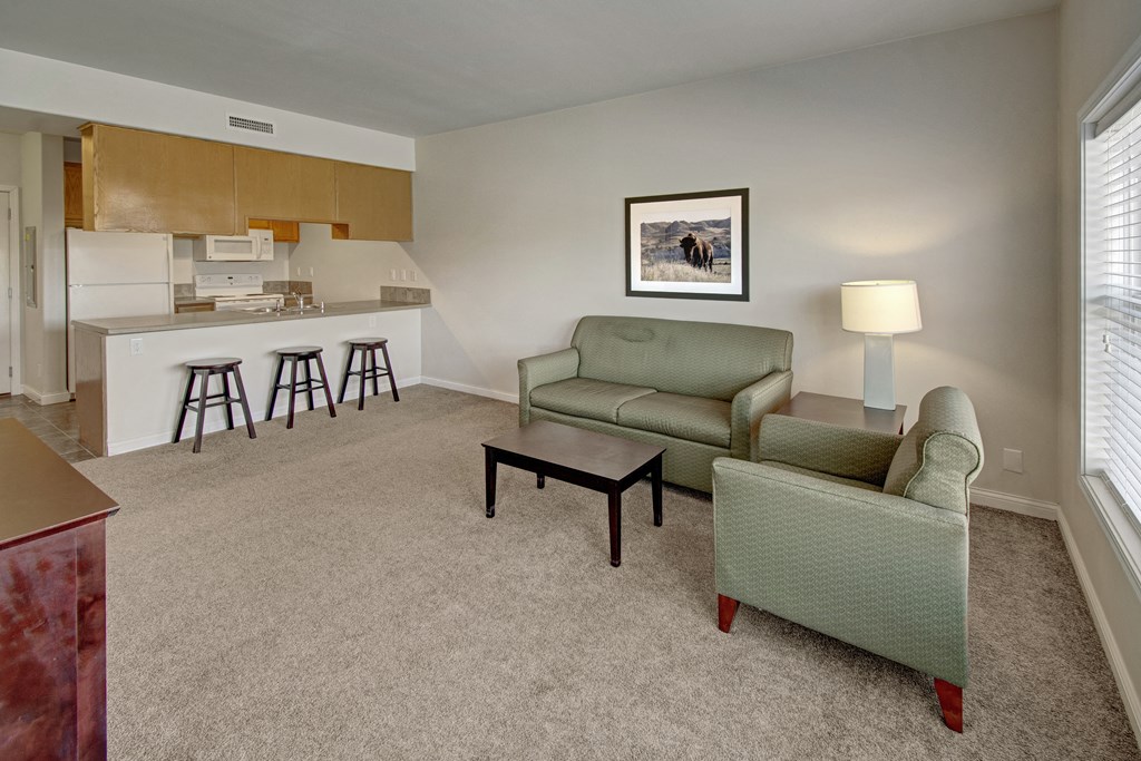 Cozy living room here at Confluence at the Harvest Hills Apartment Homes with green sofa and armchair, small coffee table, lamp, and framed bison picture. Open kitchen with three bar stools. Carpeted floor.