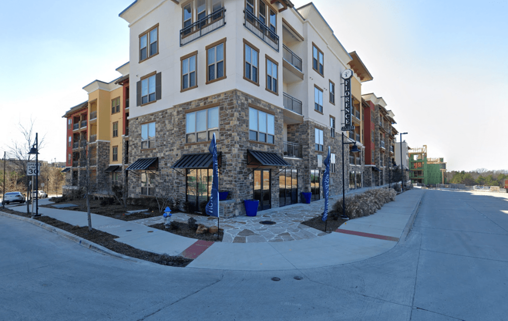 Modern Florence at the Harbor Apartment Homes leasing office with stone accents and large windows on a sunny day. It features balconies, blue planters, and a flag reading "Florence." Quiet street.