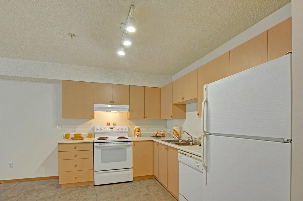 Bright kitchen here at Aspen Terrace Apartment Homes with light wood cabinets, white appliances including a refrigerator, stove, and dishwasher. Warm lighting and neutral tones create a cozy atmosphere.