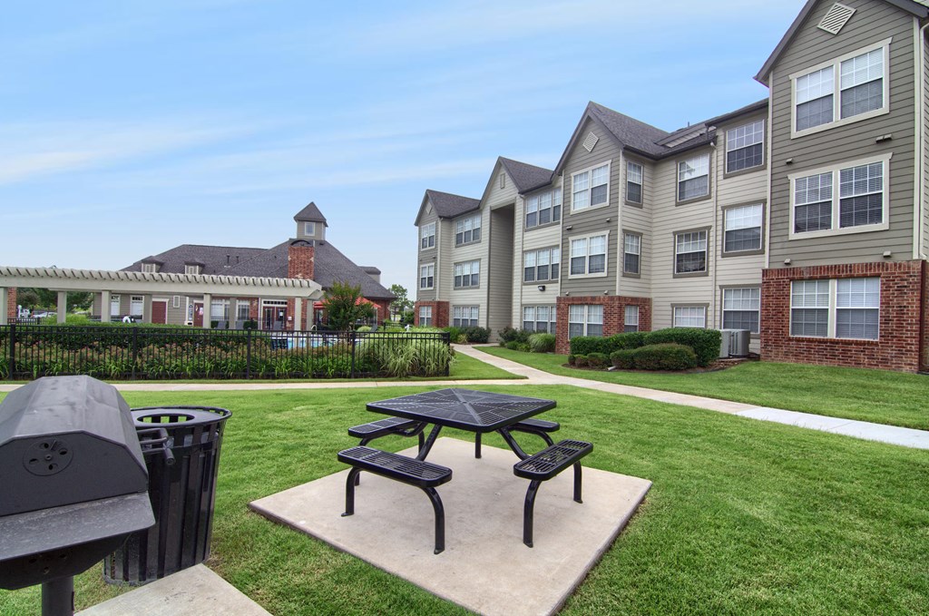 A peaceful residential courtyard here at Villas at Preston Lakes Apartment Homes with a picnic table, barbecue grill, and lush grass. Brick and beige siding apartment buildings surround the area.