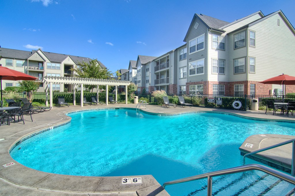 Outdoor swimming pool here at Villas at Preston Lakes Apartment Homes with clear blue water, surrounded by lounge chairs and tables with red umbrellas. Apartment buildings are in the background under a clear sky.