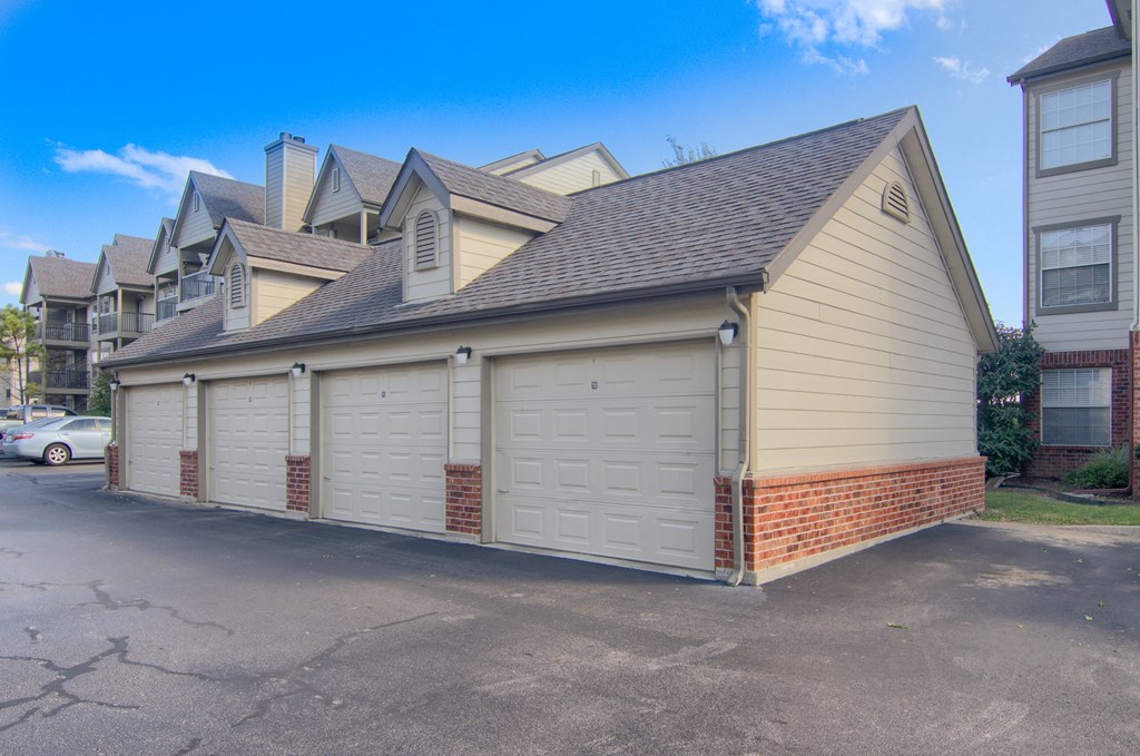 A row of beige garages here at Villas at Preston Lakes Apartment Homes, red brick accents, and gabled roofs. Clear blue sky and parked cars enhance the calm suburban setting.