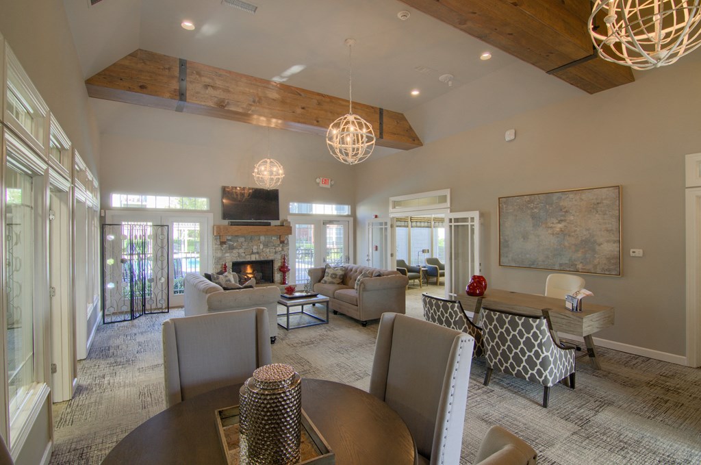 Spacious living room here at Villas at Preston Lakes Apartment Homes with vaulted ceilings and wooden beams. Features include a stone fireplace, cozy sofas, modern lighting, and a large abstract painting.