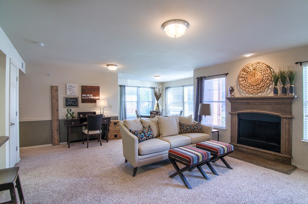 Modern living room here at Villas at Preston Lakes Apartment Homes with a cozy beige sofa, colorful cushions, striped stools, a dark wood fireplace, wall art, and large windows, creating a warm, inviting vibe.