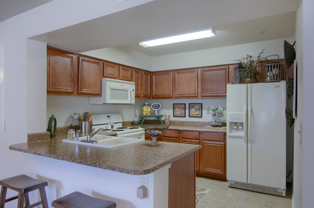 Warm kitchen here at Villas at Preston Lakes Apartment Homes with brown cabinets, white appliances, and a granite countertop. Decor includes framed art and a plant, creating a cozy atmosphere.