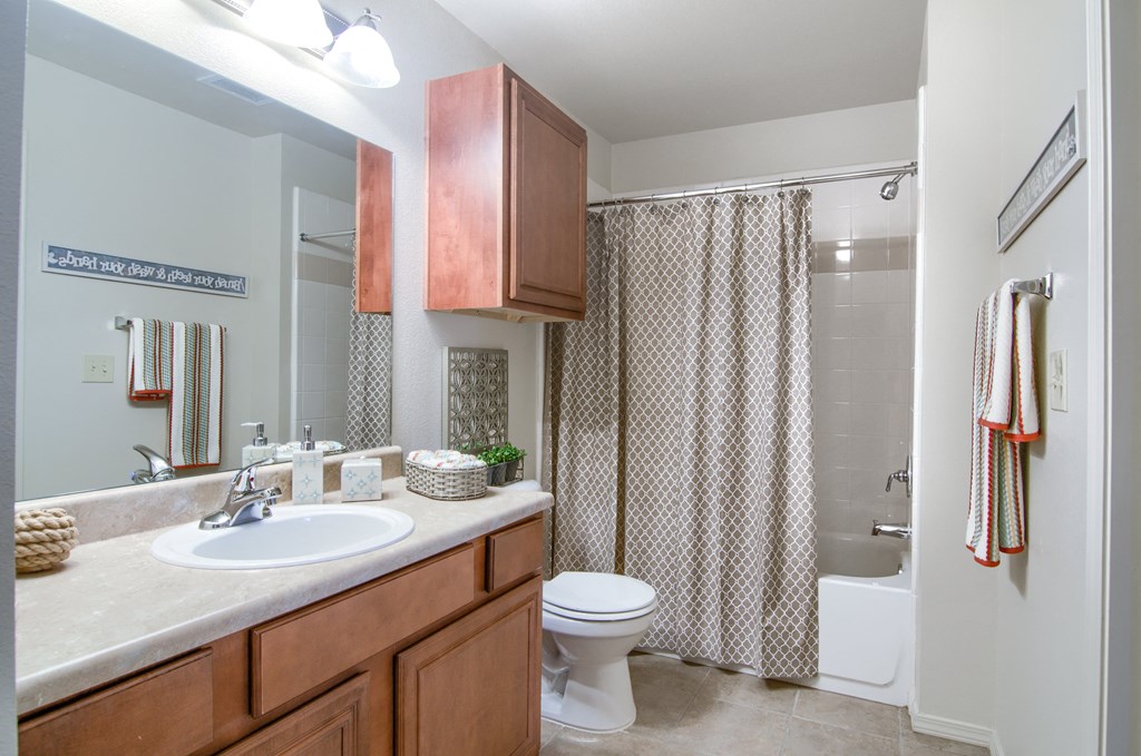 Modern bathroom here at Villas at Preston Lakes Apartment Homes with a large mirror, wooden cabinets, and a beige countertop. A patterned shower curtain and colorful striped towels add warmth.