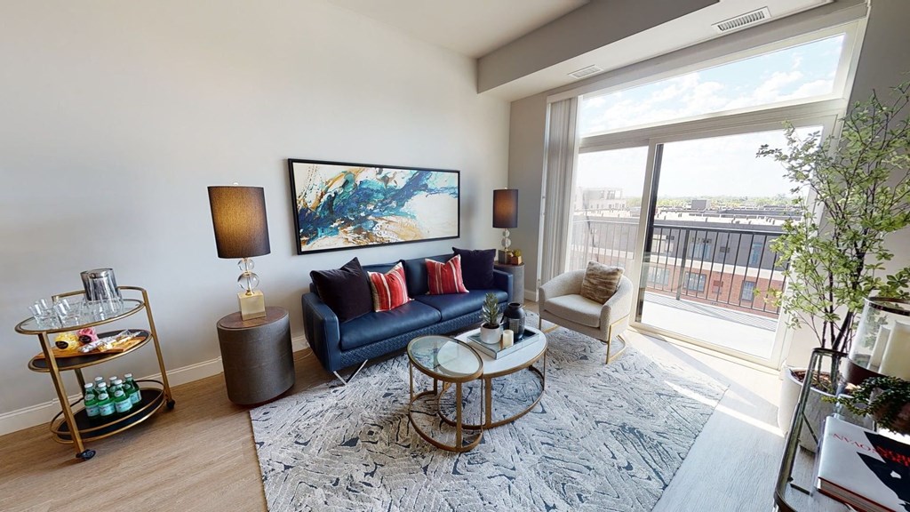 Cozy modern living room here at Daymark Uptown Apartment Homes with large window. Blue sofa, colorful cushions, abstract art, and layered coffee tables. Bright, airy atmosphere.