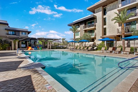 Modern Escape Apartments courtyard with a tranquil swimming pool, surrounded by palm trees and lounge chairs. Blue umbrellas offer shade under a bright sky.