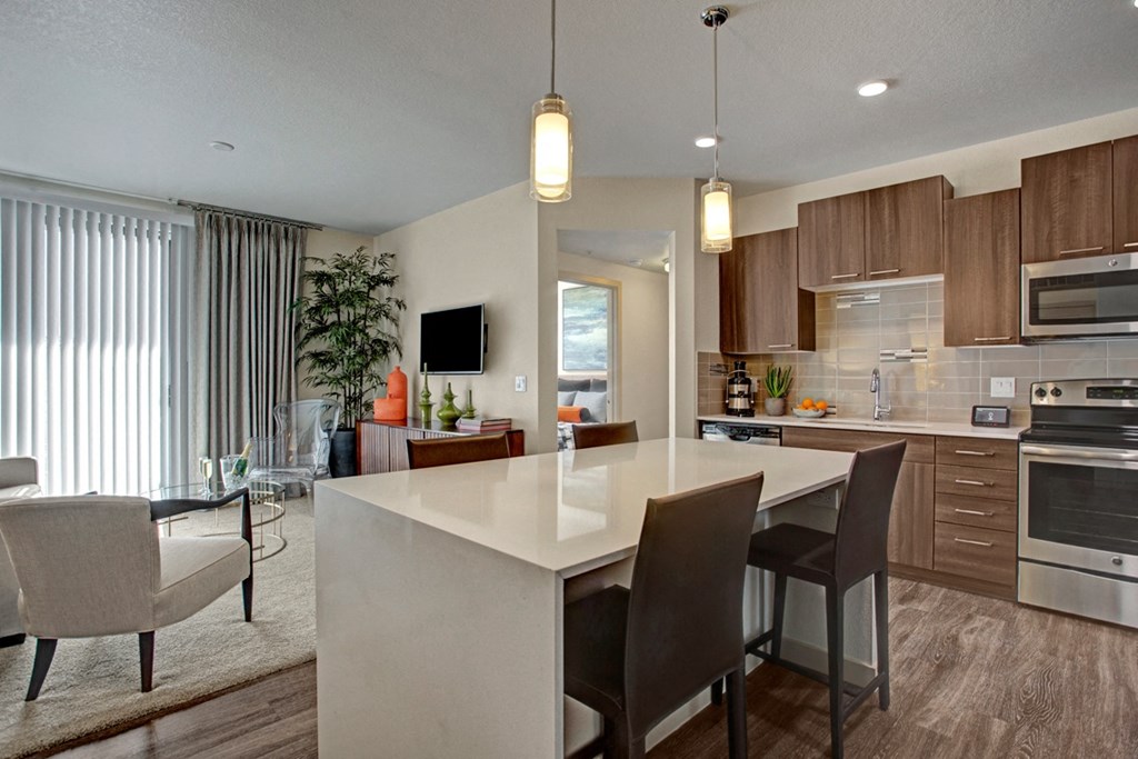 Modern kitchen and living area here at Escape Apartments with brown cabinets, stainless steel appliances, a white island with dark chairs, and cozy seating. Elegant and inviting ambiance.