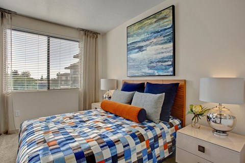 Modern bedroom here at Escape Apartments with a colorful patchwork bedspread, navy and gray pillows, and an orange bolster. Abstract wall art and a silver lamp add elegance.