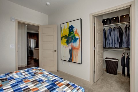 Bedroom here at Escape Apartments with abstract art and open closet. Closet contains neatly arranged blue shirts. Bedspread features a colorful geometric pattern. Neutral tones create a calm atmosphere.