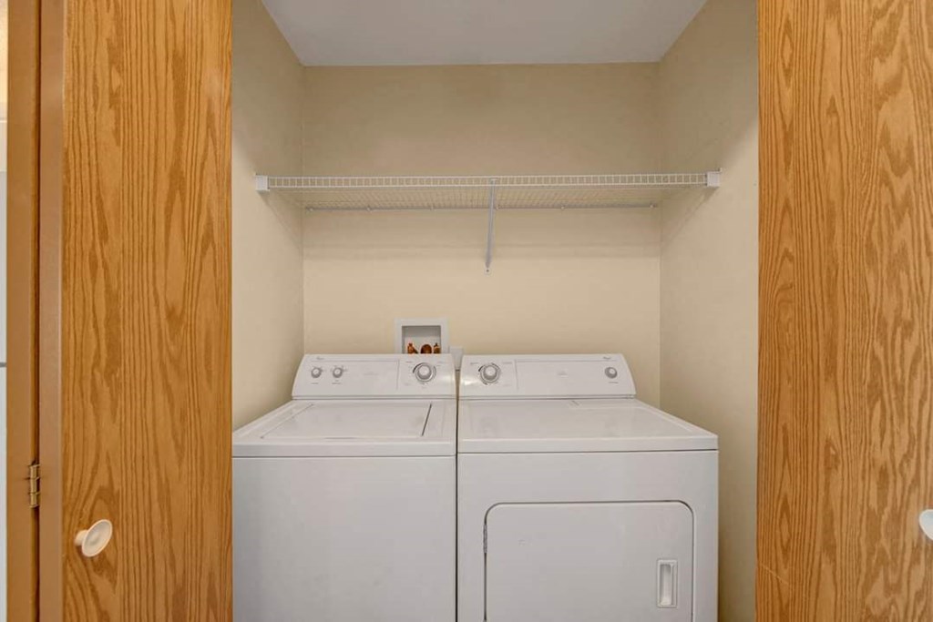 Laundry nook here at Aspen Terrace Apartment Homes with a white washer and dryer, enclosed by wooden bifold doors. Above, a wire shelf is empty. Beige walls create a tidy, minimalist vibe.